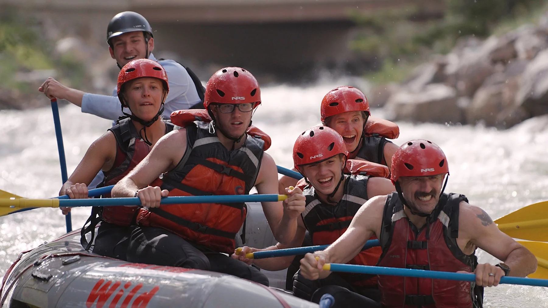 Smiling group of rafters in red helmets paddling through white water rapids on Clear Creek near Idaho Springs, Colorado, with a Mild to Wild guide.