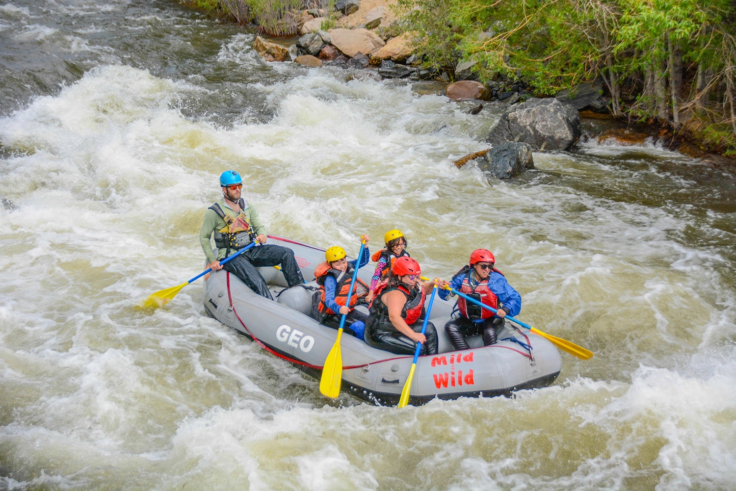 Smiling group of rafters in red helmets paddling through white water rapids on Clear Creek near Idaho Springs, Colorado, with a Mild to Wild guide.