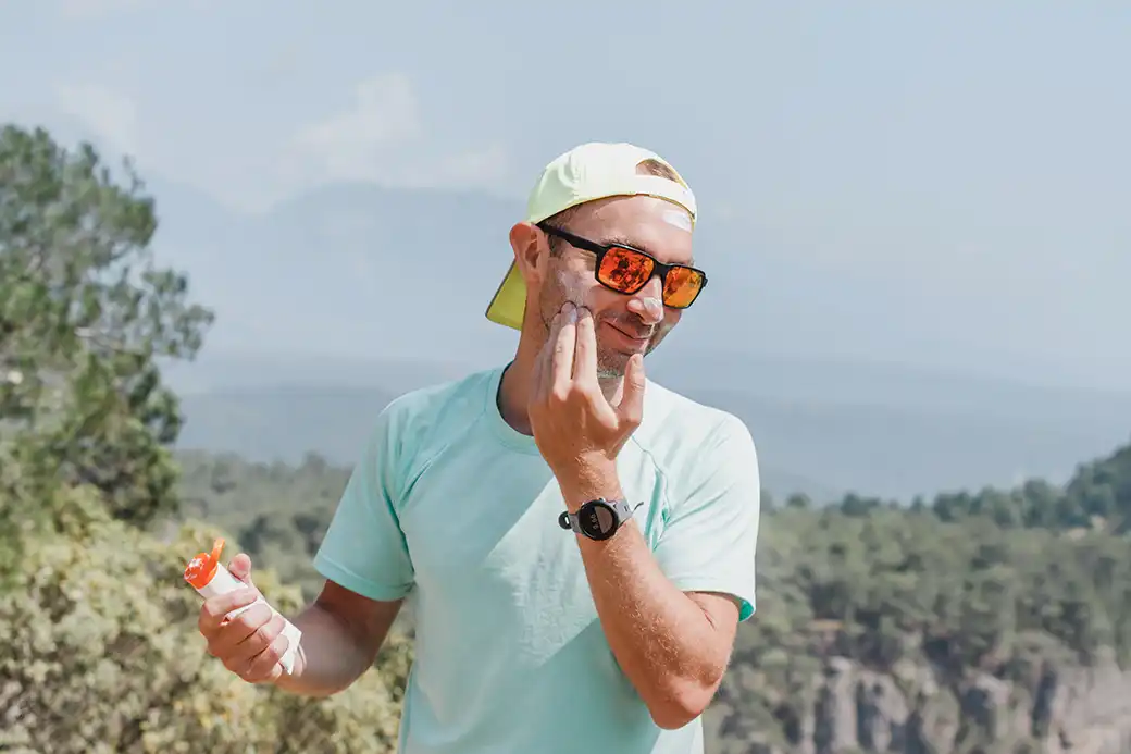 man applying sunscreen to his face while wearing a backward baseball cap and sunglasses